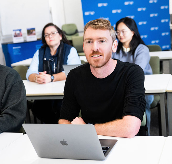 A male student sitting at a desk with a laptop and two female students sitting in the row behind him
