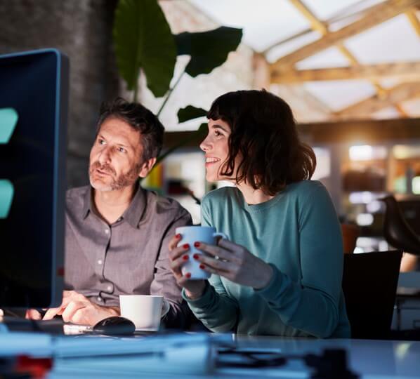 A woman holding a cup and a man look at the computer screen to the left of frame.