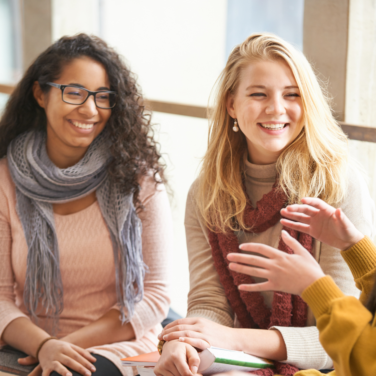 Three women sit smiling. One profile with hands up, the other two smiling and looking at third woman.