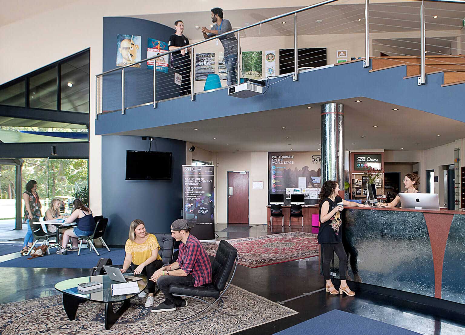 SAE Byron Bay SAE Byron Bay campus foyer. Mezzanine, front desk and tables and chairs all feature people talking and chatting.