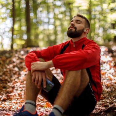 Referrals Appointments Fees Man sits on the forest ground - tree leaves blanket the forest floor. He is wearing track top, shorts and running shoes. His eyes are closed and he is leaning his back and his elbows lock around his knees. He is wearing ear buds and holding a mobile phone.