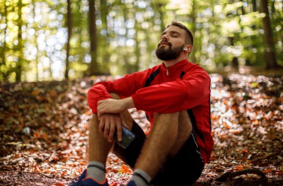Man sits on the forest ground - tree leaves blanket the forest floor. He is wearing track top, shorts and running shoes. His eyes are closed and he is leaning his back and his elbows lock around his knees. He is wearing ear buds and holding a mobile phone.