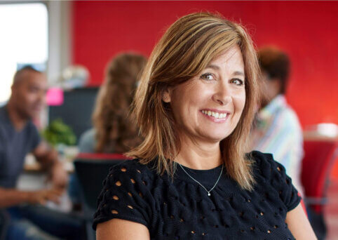 A woman smiling to camera on the right hand side of frame, with three people sitting around a desk in an office in the background | Master of Business Administration