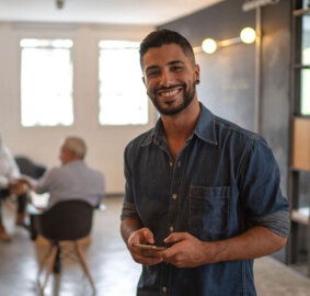 Graduate Diploma of Business Administration A man looking to camera with a phone in his hands in the foreground and a group of three people sitting in a circle on chairs in the background