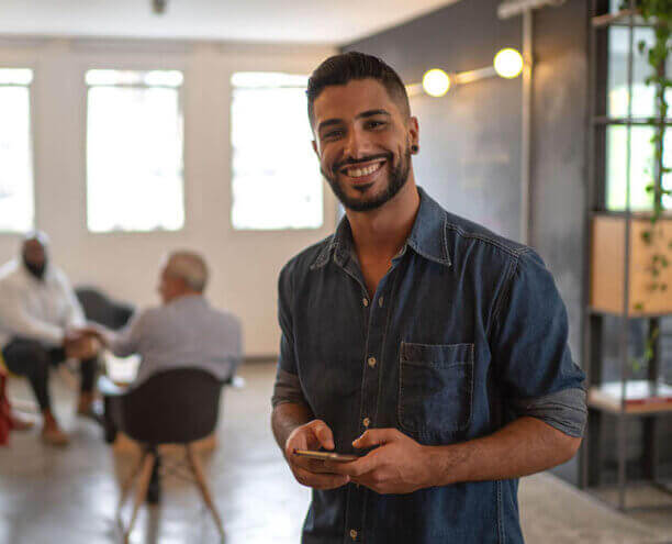 Graduate Diploma of Business Administration A man looking to camera with a phone in his hands in the foreground and a group of three people sitting in a circle on chairs in the background