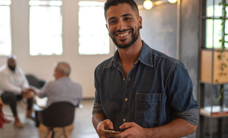 Graduate Diploma of Business Administration A man looking to camera with a phone in his hands in the foreground and a group of three people sitting in a circle on chairs in the background