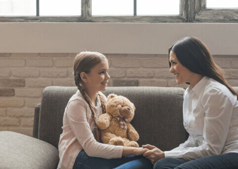 Diploma of Counselling Skills A woman on a couch sits smiles at a young girl also on the couch, holding a teddy bear. She holds her hand as they face each other.