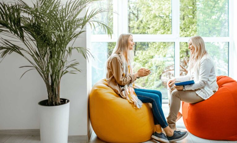 Bachelor of Psychological Science (Honours) Two people sit on bean bags in front of large wall of windows. They smile at each other. One person holds a clipboard and pen. A large potted plant is in the foreground and to the left of people.