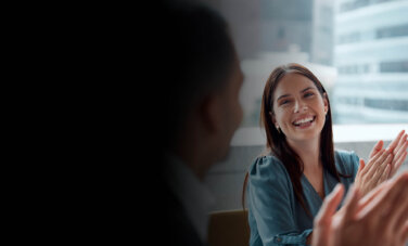 Woman sitting at a board room with city scape in background, smiles and claps looking at man also clapping - but blurry and obscured in foreground.