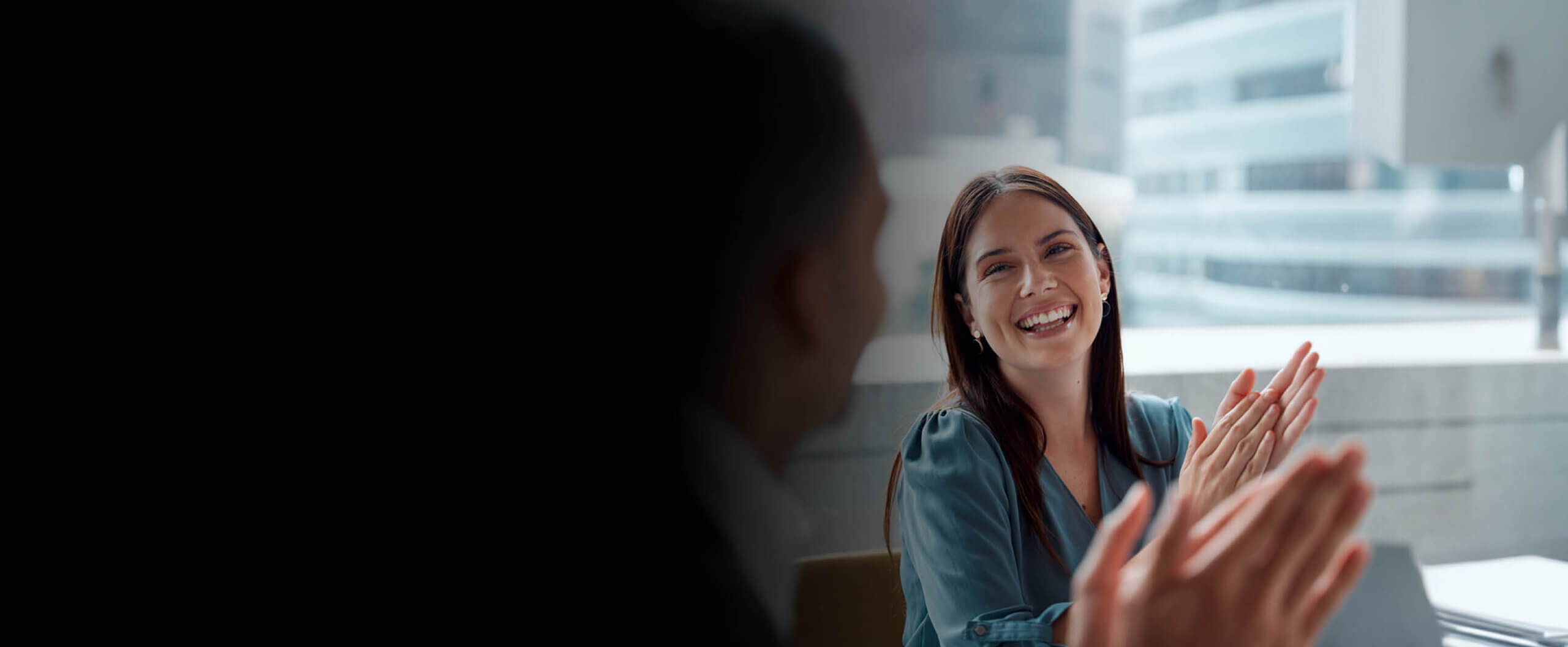 Woman sitting at a board room with city scape in background, smiles and claps looking at man also clapping - but blurry and obscured in foreground.