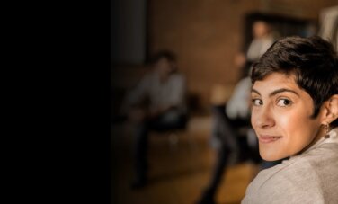 Woman with short cropped hair and padlock earring looks over her shoulder to camera. The background is a group of people in circle of chairs. They are blurry and nondescript.