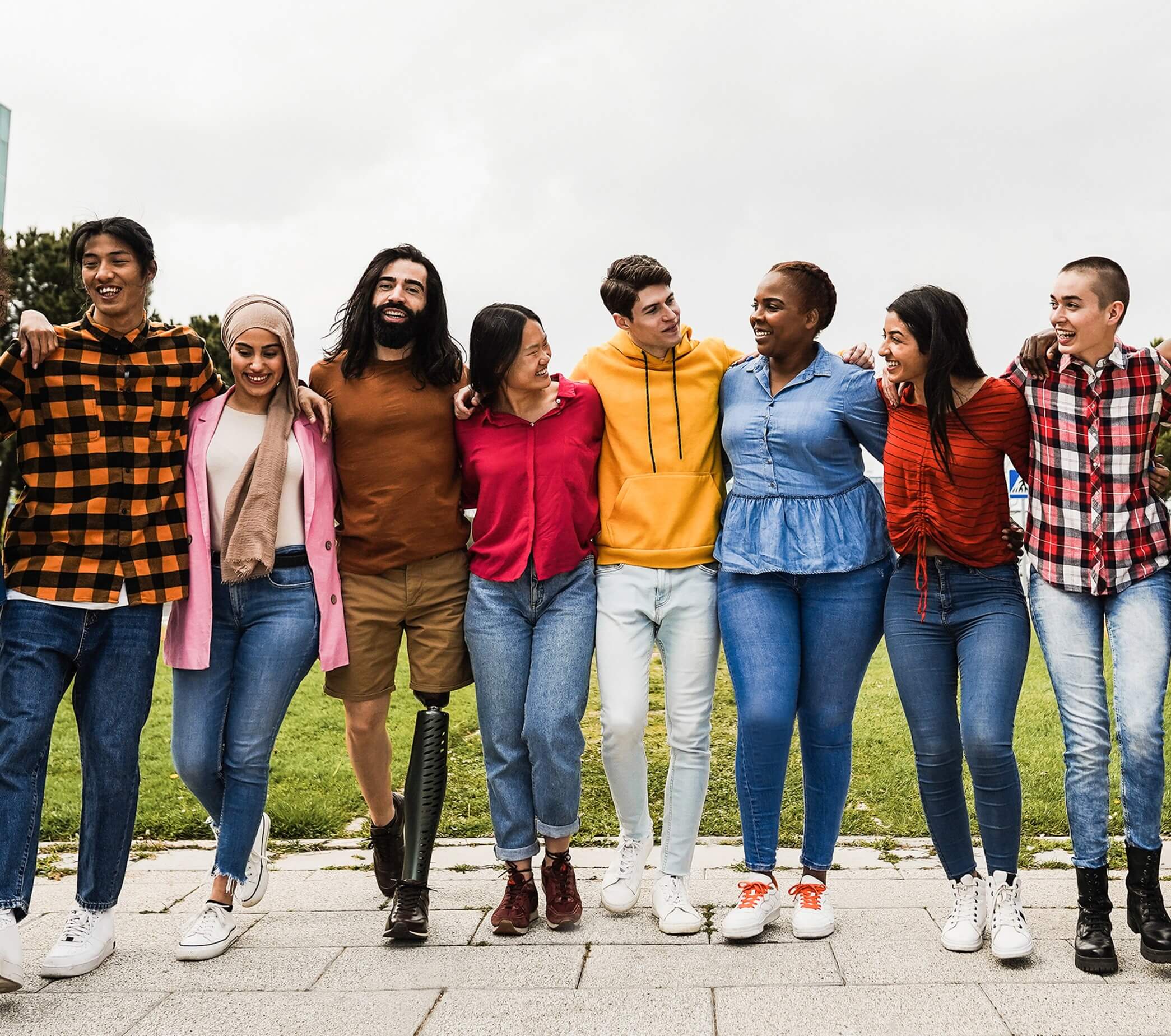 A group of people stand, arm in arm. They all appear young and of mixed ethnicity. They are standing outside, with trees and builds to the left and right of them. They all look at each other, smiling and laughing.