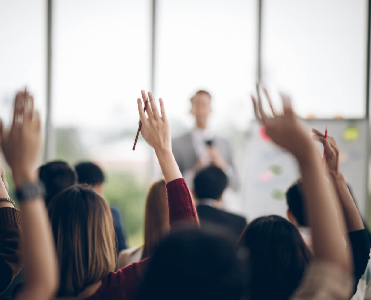 Back of people's heads and raised hands at corporate presentation with speaker and whiteboard out of focus in background Rear view of heads and raised hands at corporate presentation with speaker and whiteboard out of focus in background ACAP Careers Culture