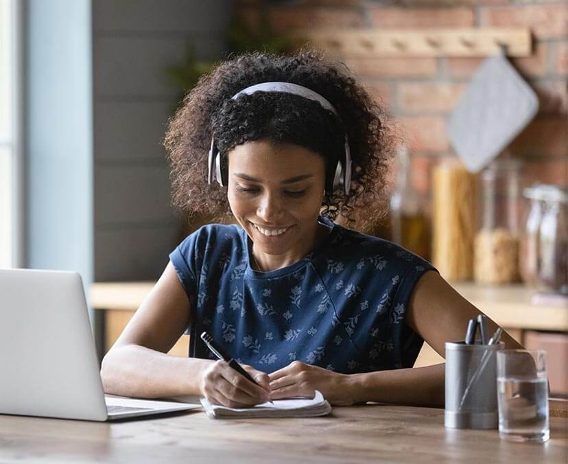 ACAP Info Night Woman with curly hair wears headphones and looks down at notepad with pen in hand. She is smiling. A laptop, pen holder and glass of water on desk in front of her. She seems to be in a kitchen - food jars of pasta blurry in background.