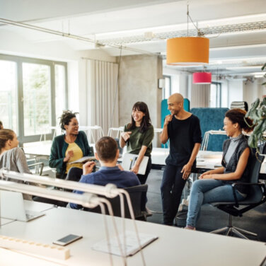 A group of people sitting and standing in a circle formation, talking in an open plan office space ACAP Careers Campus Operations