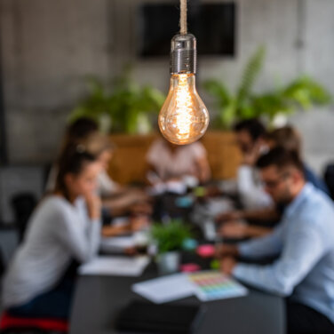 Out of focus view of office workers sitting around a desk with a lightbulb in the foreground ACAP Careers Management