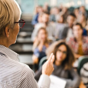 Mature female lecturer with short blonde hair standing in front of a class with two fingers pointed in the air ACAP Careers Psychology