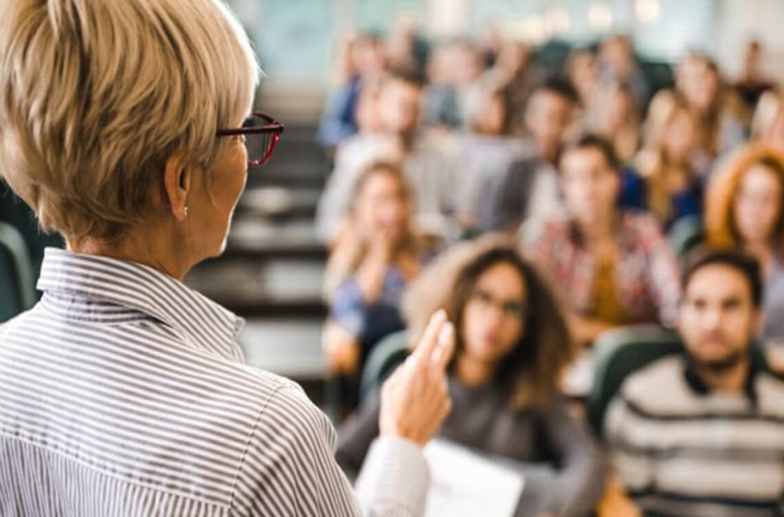 Mature female lecturer with short blonde hair standing in front of a class with two fingers pointed in the air ACAP Careers Psychology