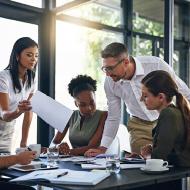 A group of office workers sitting and standing around a desk viewing documents ACAP Careers Systems, Finance & Human Resources