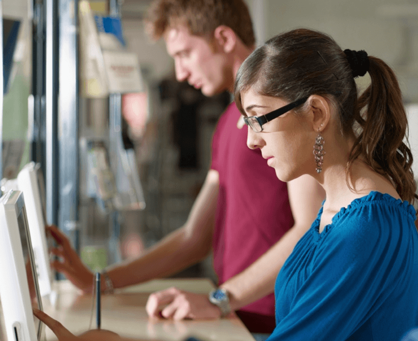 Side view of two students standing at a desk working on desktop computers,