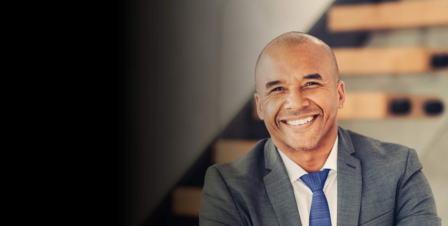 A man with a shaved head smiling towards camera, wearing a dark grey suit and blue tie - ACAP Graduate Certificate of Professional Accounting (2880x1188)