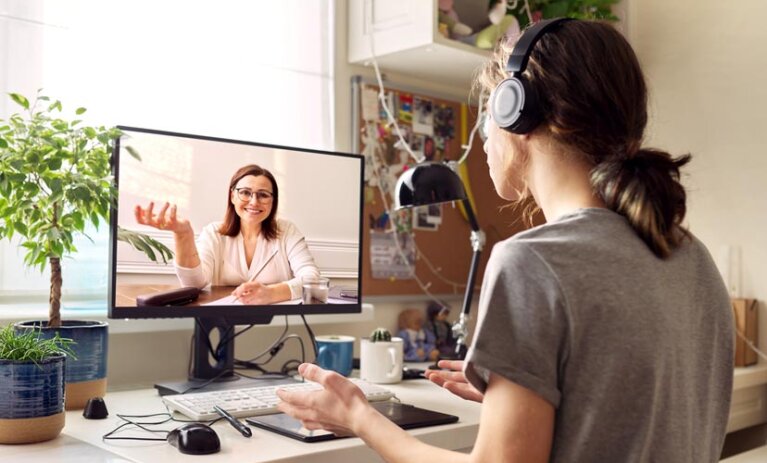 A virtual video call with a woman on a monitor screen. The monitor is on an office desk, in a home, with another person sitting in office chair with headphones.