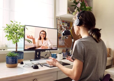 Woman sits in front of computer monitor with headphones on. She is sitting in an home office and the monitor displays an image of someone speaking to camera. Like a Zoom or virtual video call.