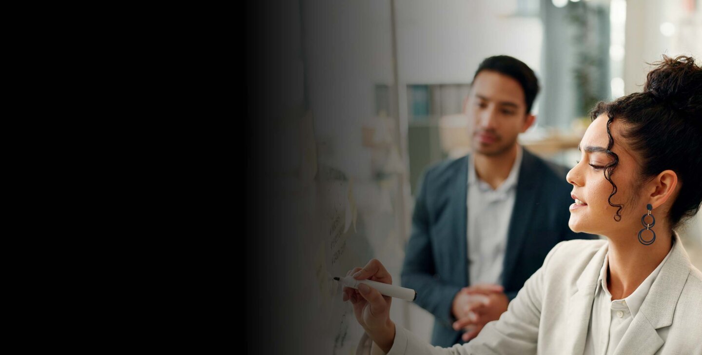 Woman stands at white board in foreground, holding a marker, speaking aloud to man in the background.