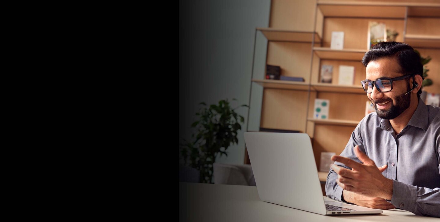 APPLIED COACHING Man wears headset and sits in front of open laptop at desk. A book shelf in background