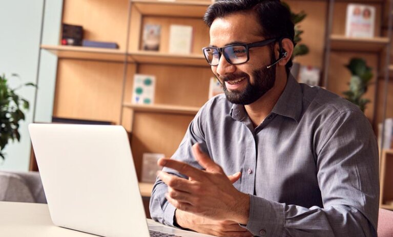 Applied Coaching Man wears headset and sits in front of open laptop at desk. A book shelf in background