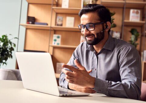 Man wears headset and sits in front of open laptop at desk. A book shelf in background