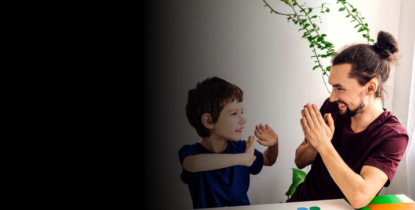 Man with top bun and tee shirt on sits at small table with hands in prayer position. He is smiling with young boy who has his hands up but looking at man.