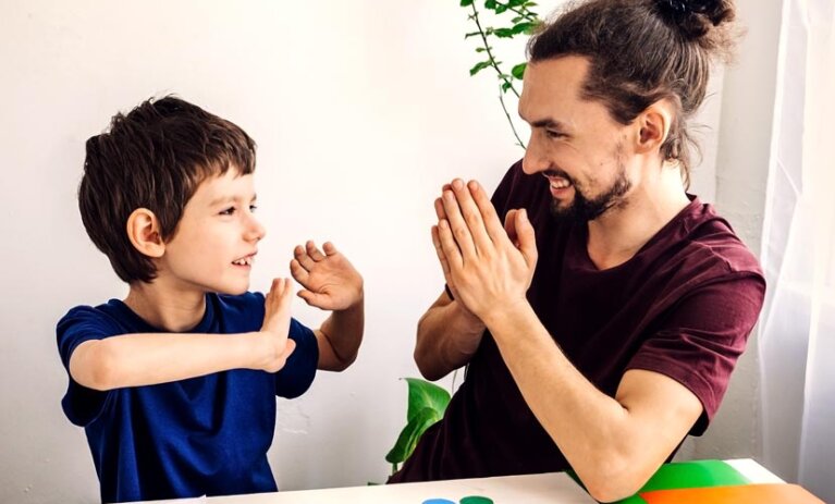 Man with top bun and tee shirt on sits at small table with hands in prayer position. The table has letters and numbers and shapes and coloured paper. He is smiling with young boy who sits opposite him, with his hands up and looking at man.