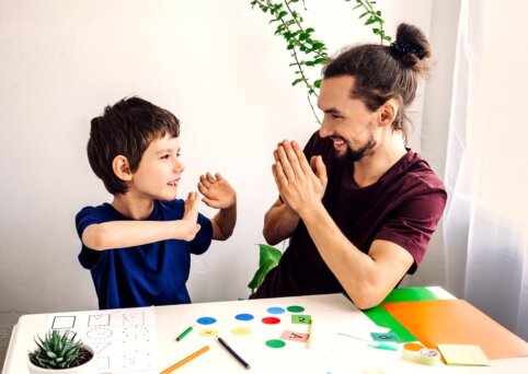 Man with top bun and tee shirt on sits at small table with hands in prayer position. The table has letters and numbers and shapes and coloured paper. He is smiling with young boy who sits opposite him, with his hands up and looking at man.