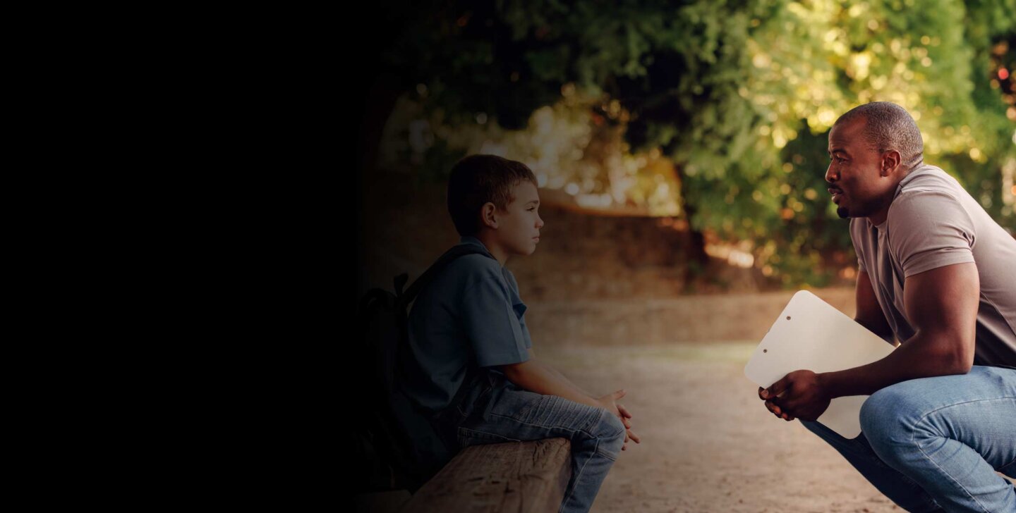 Man crouches down to speak to small child sitting on a log. Both are outdoors. Man holds a clipboard.