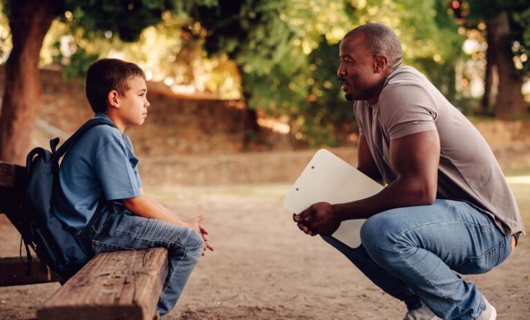 Man crouches holding a clipboard. He is speaking to small child, who is wearing a backpack, and sitting on a wooden bench. Both are outdoors.