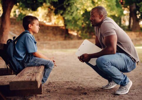 Man crouches holding a clipboard. He is speaking to small child, who is wearing a backpack, and sitting on a wooden bench. Both are outdoors.
