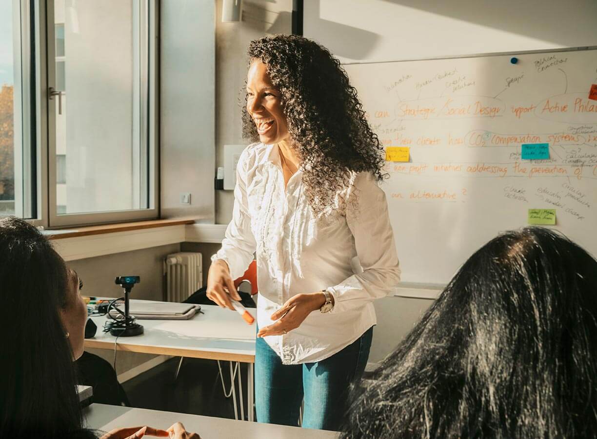 Smiling teacher standing in front of a class with a whiteboard behind them and a whiteboard marker in their hand