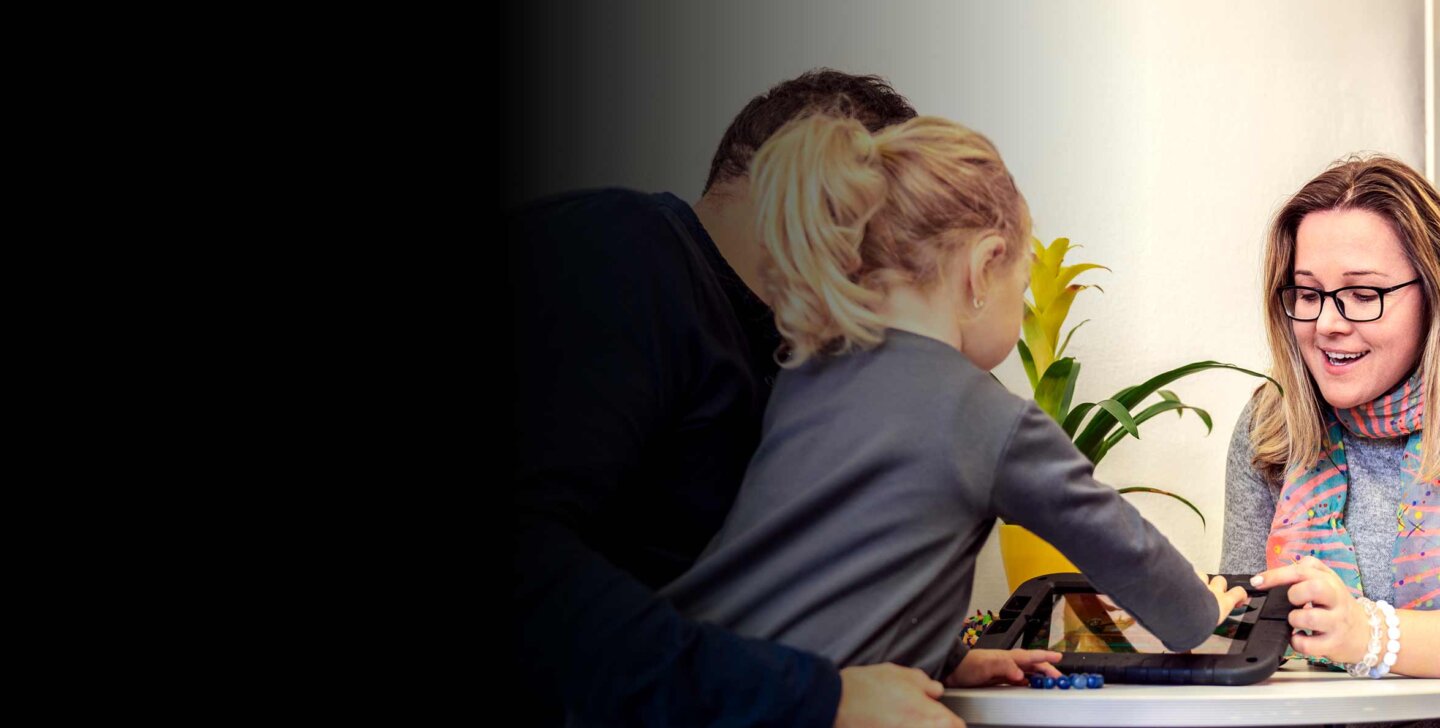 Young child sits on mans lap. Uses the touch screen of a tablet. Woman sits opposite pointing and speaking about the screen.