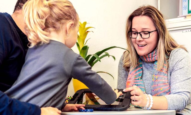 Woman sits at table, pointing and speaking about a tablet screen as young child uses the touch screen. Child is sitting on mans lap (man faces away from camera and hidden behind child).