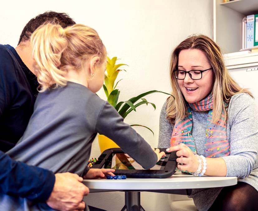 Master of Counselling and Psychotherapy A young child sitting on a man's lap and touching the screen of a tablet, sitting across a table from a female counsellor | Master of Counselling and Psychotherapy