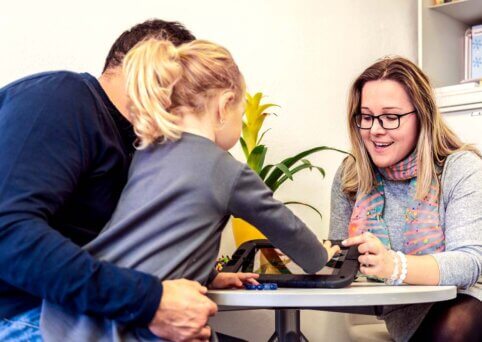 Man holds child on lap - his face obscured by child. Young girl also faces away from camera but is leaning forward to use touchscreen of tablet on a table. Woman sits across from father/daughter at the table, pointing and speaking about a tablet screen.