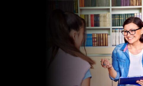 Woman sits in front of shelves of books. She has a clipboard and a pen and she is smiling at a woman (back to camera & foreground).