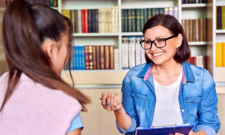 Woman sits in front of bookshelf. She has a clipboard and she is smiling at a woman (back to camera).