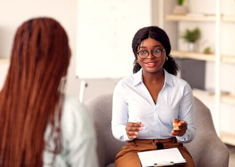 Woman sits on a seat with a clipboard on her lap. She sits in front of a woman with long hair (back to camera). They are in an office environment with a bookshelf in background.