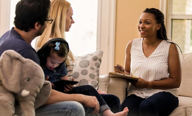 A couple sits on a couch. A young child on a tablet is on the mans lap. A woman sits opposite on another couch. She is speaking and smiling and holds a clipboard and pen in her hand.