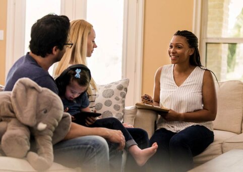 A couple sits on a couch. A young child on a tablet is on the mans lap. A woman sits opposite on another couch. She is speaking and smiling and holds a clipboard and pen in her hand.