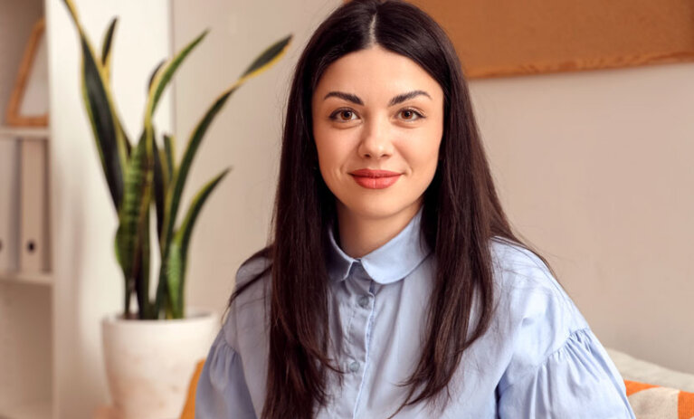 Woman sits on couch and looks to camera. She is sitting on a couch and a plant and bookshelf is in the background.