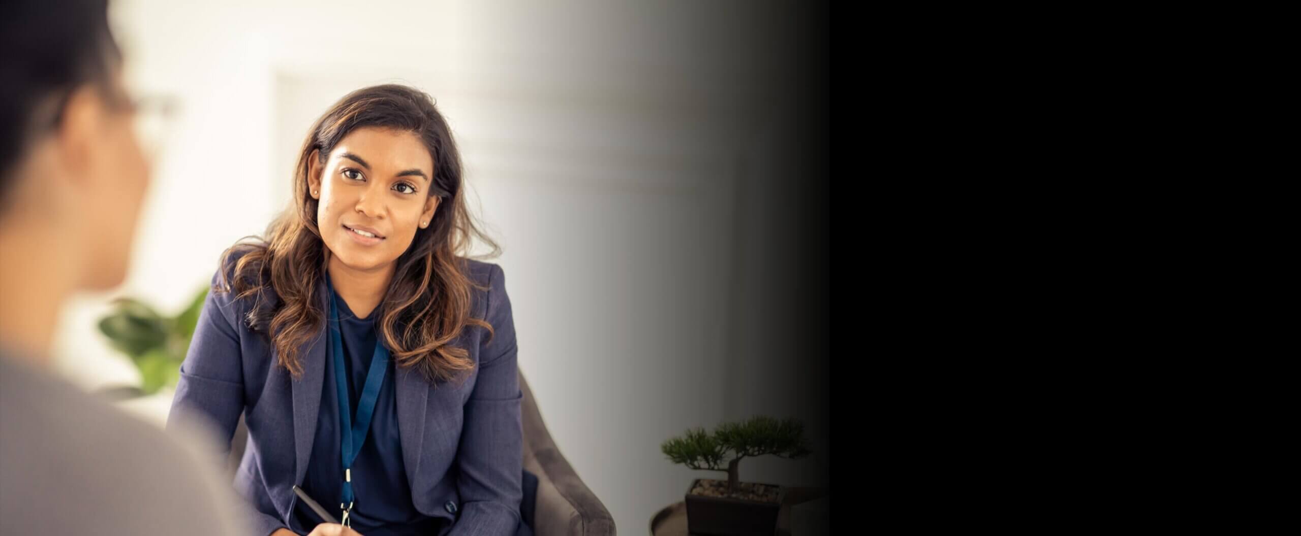 Woman sitting on a chair and facing towards camera with tablet and stylus in hand, looking across at another person in the foreground
