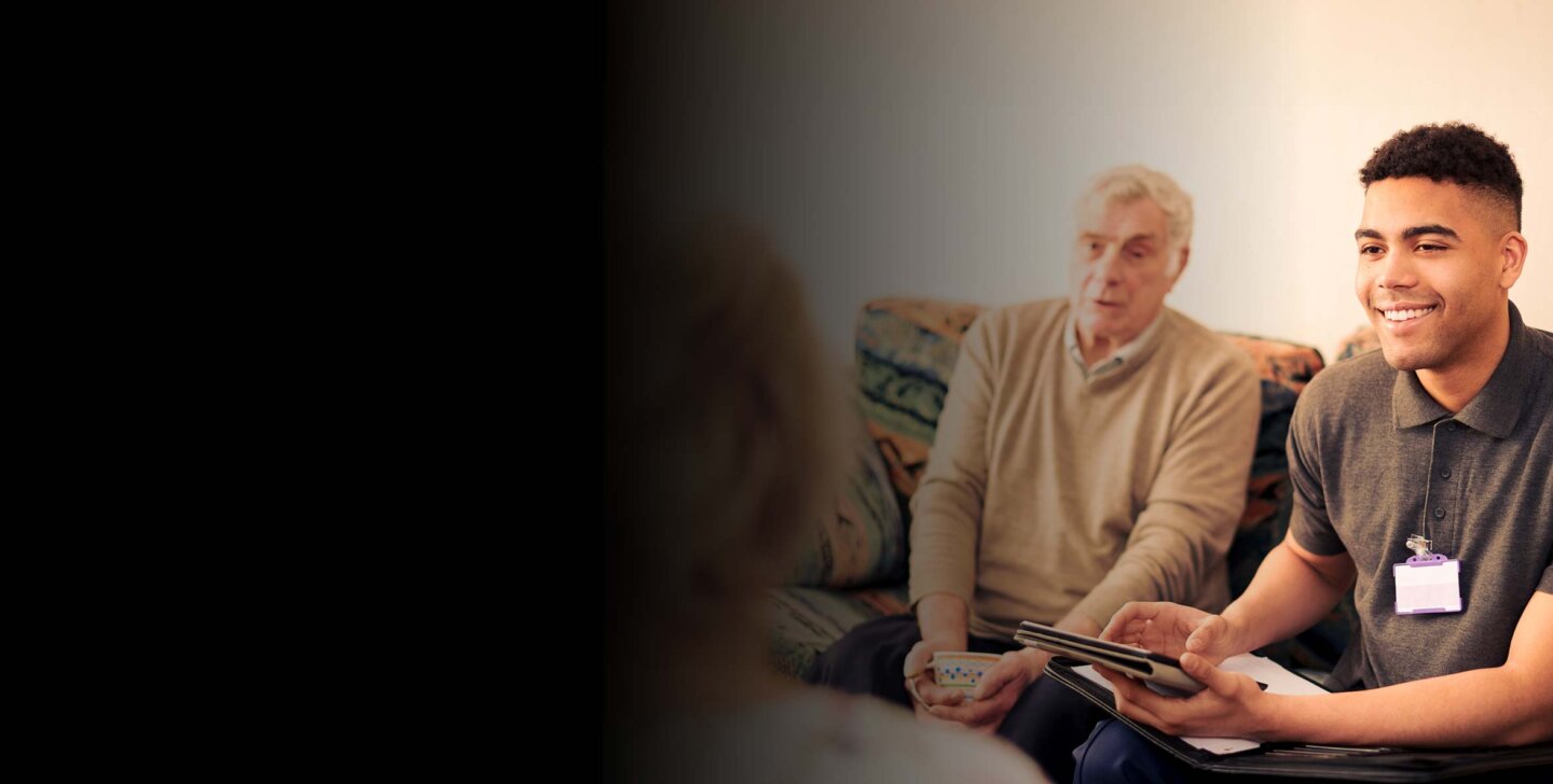 Young social worker sitting on the couch with an older man, with tablet in hand